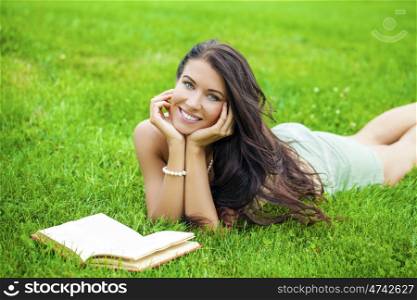 Young beautiful brunette girl with book lying on the green grass in the summer park