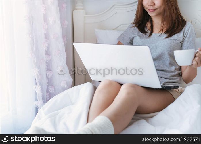 Young beautiful brunette hair woman using computer laptop and drinking coffee while sitting on the bed in the morning. close up.