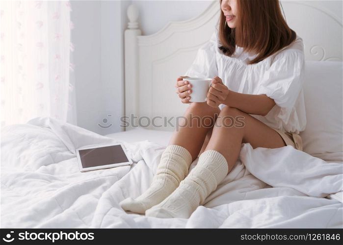 Young beautiful brunette hair woman with a cup of coffee sitting down on the bed by the window. close up, copy space Lazy day off concept