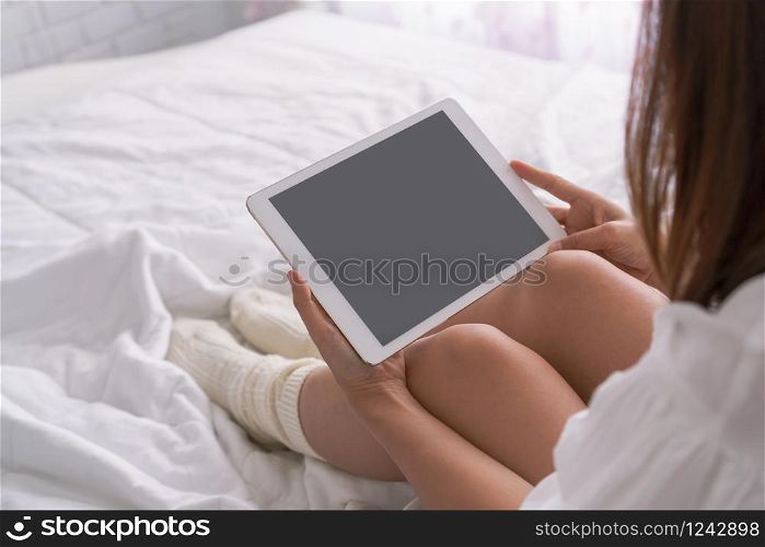 Young beautiful brunette hair woman with tablet computer sitting down on the bed by the window. Top view, copy space Lazy day off concept