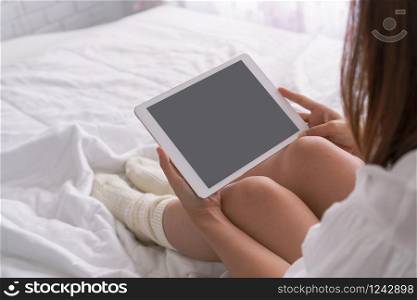 Young beautiful brunette hair woman with tablet computer sitting down on the bed by the window. Top view, copy space Lazy day off concept