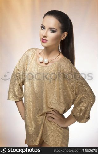 Young, beautiful brunette in sparkling gold dress and beautiful, big, gold necklace. She has long, straight hair clipped in ponytail. She is wearing purple make up and looking at camera.