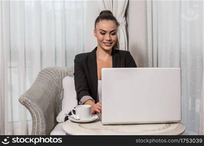 Young beautiful business woman browsing in laptop while sitting at table and drinking tea in hotel room. Smiling female working at laptop in hotel