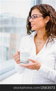Young beautiful businesswoman taking break, standing in front of office window, holding coffee cup, looking away, smiling.