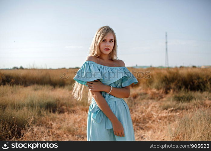 young beautiful caucasian blonde girl in light blue dress stands on the field with the sun-scorched grass