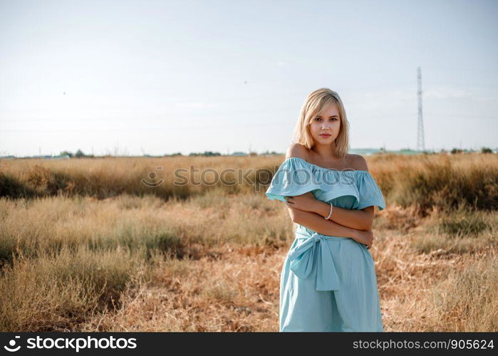 young beautiful caucasian blonde girl in light blue dress stands on the field with the sun-scorched grass