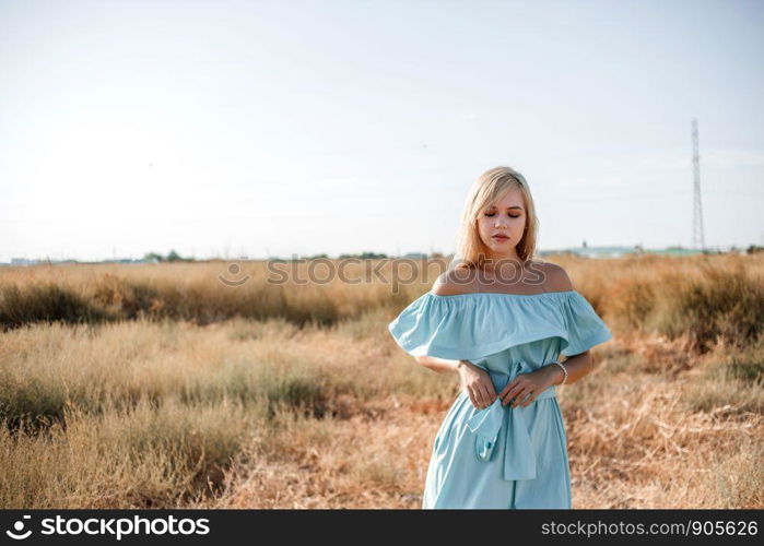 young beautiful caucasian blonde girl in light blue dress stands on the field with the sun-scorched grass