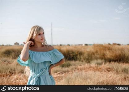 young beautiful caucasian blonde girl in light blue dress stands on the field with the sun-scorched grass