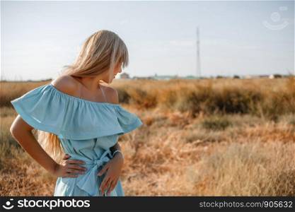 young beautiful caucasian blonde girl in light blue dress stands on the field with the sun-scorched grass