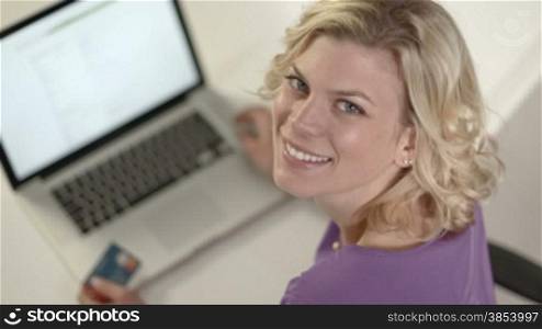 Young beautiful caucasian blonde woman buying on internet with laptop pc and credit card and looking over shoulders at camera. High angle view