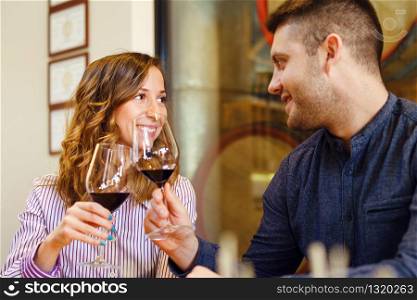 Young beautiful caucasian couple in love toasting with glasses of red wine while sitting by the table at home or restaurant man and woman girl looking each other in the eyes smiling happy flirting
