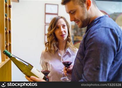 Young beautiful caucasian woman girl looking to her boyfriend man while he is picking a wine bottle from the shelf reading holding glasses of red wine couple at winery or home husband and wife