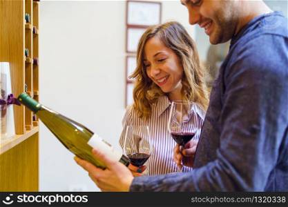 Young beautiful caucasian woman girl smiling while her boyfriend man is picking a wine bottle from the shelf reading holding glasses of red wine couple at winery or home husband and wife having fun