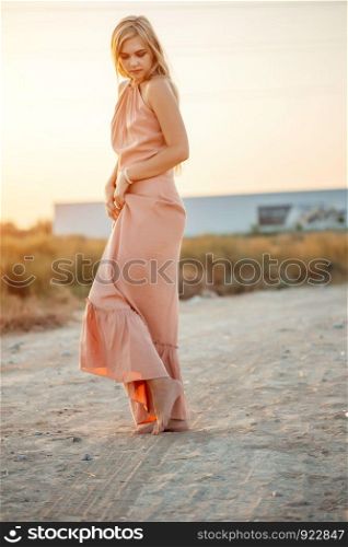 young beautiful Caucasian woman in pink dress walks on sand with bare feet during sunset