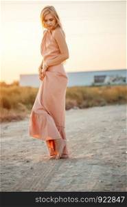 young beautiful Caucasian woman in pink dress walks on sand with bare feet during sunset