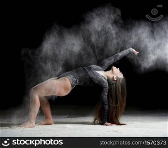 young beautiful caucasian woman with long hair is dressed in a sports black bodysuit stands in a bent pose and throws white flour, black background, gymnastic exercises