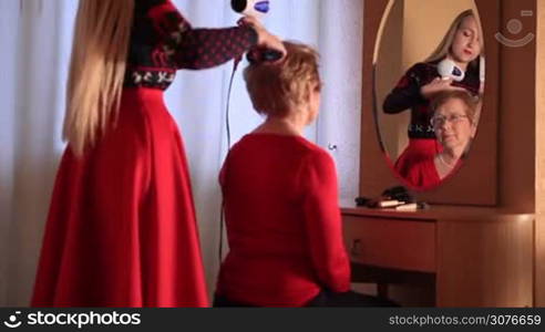 Young beautiful daugther holding haidryer and doing mother&acute;s hair with love at home near the mirror. They&acute;re preparing for family Christmas celebration.