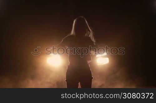 Young beautiful excited woman listening music and dancing with long blond hair flying away in bright car headlights in night time. Overjoyed female in shorts spending leisure dancing in clouds of smoke over yellow car headlights at night. Slow motion