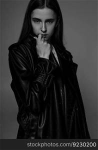 Young beautiful fashion model in long leather coat and dark jeans on dark grey.