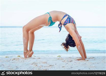 Young beautiful fit caucasian woman in summer girl bikini swimsuit practice yoga at the beach by the sea or ocean in sunny day stretching on the vacation bridge position