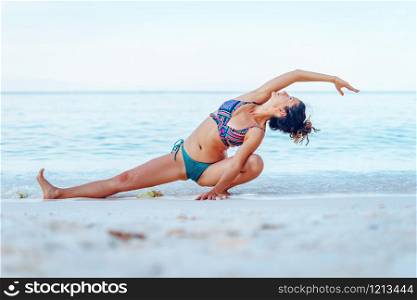 Young beautiful fit caucasian woman in summer girl bikini swimsuit practice yoga at the beach by the sea or ocean in sunny day stretching on the vacation