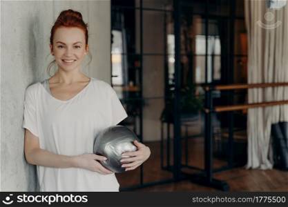 Young beautiful fit woman with smile with mini size fitball looking positively at camera while leaning on grey concrete wall in fitness center ambience, female resting after intensive pilates class. Happy active ginger woman with mini fitball relaxing after workout in fitness studio