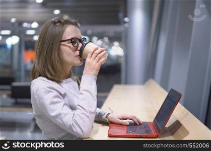 Young, beautiful girl drinks coffee standing at the table with laptop at the airport.. Young, beautiful girl drinks coffee standing at the table with laptop at the airport