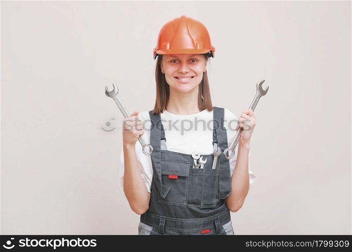 Young beautiful girl engineer in a gray uniform and protective helmet smiles with tools, wrenches in her hands. Equality between men and women. Repairwoman posing on a white background. Young beautiful girl engineer in a gray uniform and protective helmet smiles with tools, wrenches in her hands. Equality between men and women. Repairwoman posing on a white background.