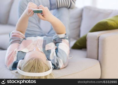 Young beautiful girl enjoying music through headphones, laying on sofa at home