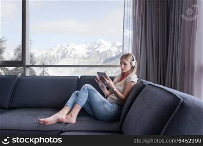 Young beautiful girl enjoying music through headphones, laying on sofa near the window at home