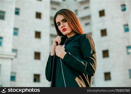 Young beautiful girl in a black jacket poses near a high white house.