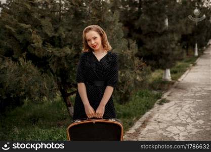 young beautiful girl in a black vintage polka dot dress with a vintage suitcase in her hands.