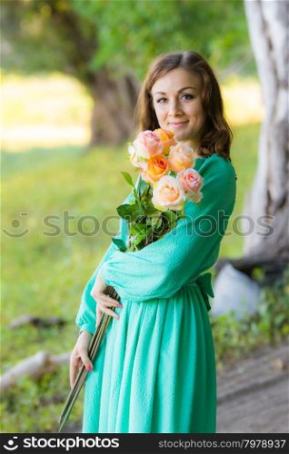 Young beautiful girl in a long dress walking in the woods on a warm summer day. Portrait of a girl with a bouquet of roses walking in the woods