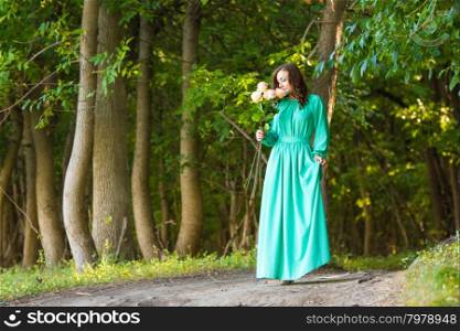 Young beautiful girl in a long dress walking in the woods on a warm summer day. A girl in a long dress walking along the road and sniffing a bouquet of roses