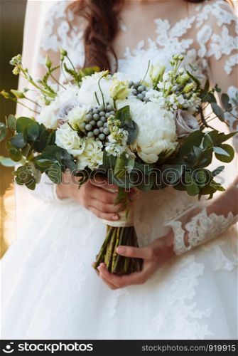 Young beautiful girl in an elegant dress is standing and holding hand bouquet of pastel flowers and greens with ribbon at nature. The bride holds a wedding bouquet outdoors