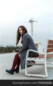 young beautiful girl in Burgundy stockings and gray coat sitting on a white bench with a camera at sunset