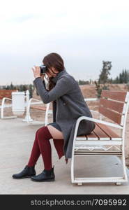 young beautiful girl in Burgundy stockings and gray coat sitting on a white bench with a camera at sunset