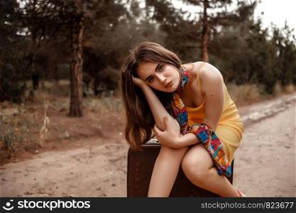 young beautiful girl in yellow dress with colorful scarf sitting on vintage suitcase on soaked muddy road after rain