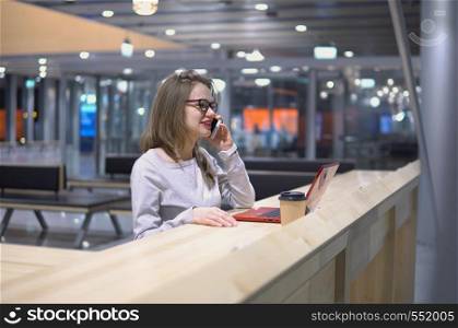 Young, beautiful girl talking on the phone standing at a table with a laptop and a cup of coffee at the airport.. Young, beautiful girl talking on the phone standing at a table with a laptop and a cup of coffee at the airport