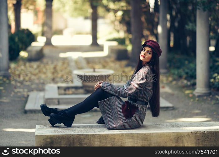 Young beautiful girl wearing winter coat and cap sitting on a bench in urban park. Lifestyle and fashion concept.. Young beautiful girl wearing winter coat and cap sitting on a bench in urban park.