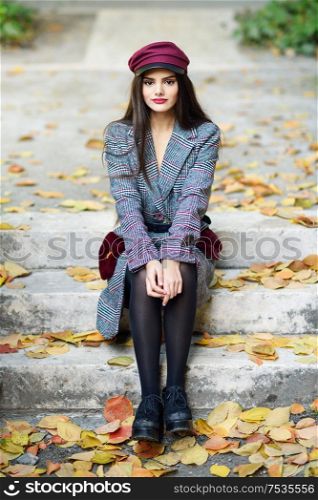Young beautiful girl wearing winter coat and cap sitting on steps full of autumn leaves Lifestyle and fashion concept.. Young beautiful girl wearing winter coat and cap sitting on steps full of autumn leaves