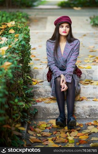 Young beautiful girl wearing winter coat and cap sitting on steps full of autumn leaves Lifestyle and fashion concept.. Young beautiful girl wearing winter coat and cap sitting on steps full of autumn leaves