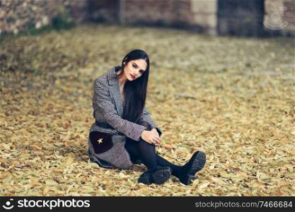 Young beautiful girl with very long hair wearing winter coat sitting on the floor of an urban park full of autumn leaves. Lifestyle and fashion concept.. Beautiful girl wearing winter coat sitting on the floor of an urban park full of autumn leaves.