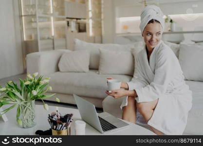 Young beautiful happy woman in bathrobe with cup of tea or coffee feeling relaxed after taking shower or bath at home, sitting on sofa in living room and looking aside while working on laptop. Young happy woman freelancer in bathrobe drinking tea and working remotely on laptop