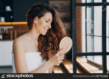 Young beautiful hispanic woman brushing long healthy brown curly hair by wooden hairbrush after shower. Female wrapped in towel do morning beauty routine at home. Haircare cosmetics advertising.. Young girl combing long healthy hair by wooden hairbrush after shower. Haircare cosmetic advertising