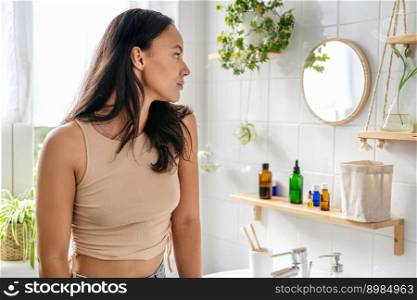 Young beautiful hispanic woman looking into mirror in bathroom. Natural cosmetics, wellness, self-care. Young beautiful hispanic woman looking into mirror in bathroom