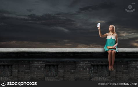 Young beautiful lady sitting on roof with book on knees and mobile phone in hands. Tradition and modernity