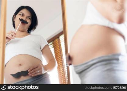 Young Beautiful Mother having fun standing looking at mirror and pretending to be Father with moustache. Happy funny Asian pregnant woman enjoying with Mustache on face and belly.