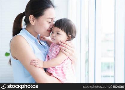 Young beautiful mother holding and kissing her cute little Caucasian 7 months newborn baby on her hands, standing near window at home. Happy family bonding, spending time together. Copy space