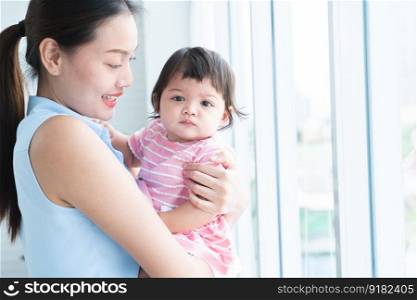 Young beautiful mother holding and looking at her cute little Caucasian 7 months newborn baby on her hands, standing near window at home. Happy family bonding, spending time together. Copy space